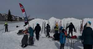 Lots of people and skis in front of a mound of snow with an event flag