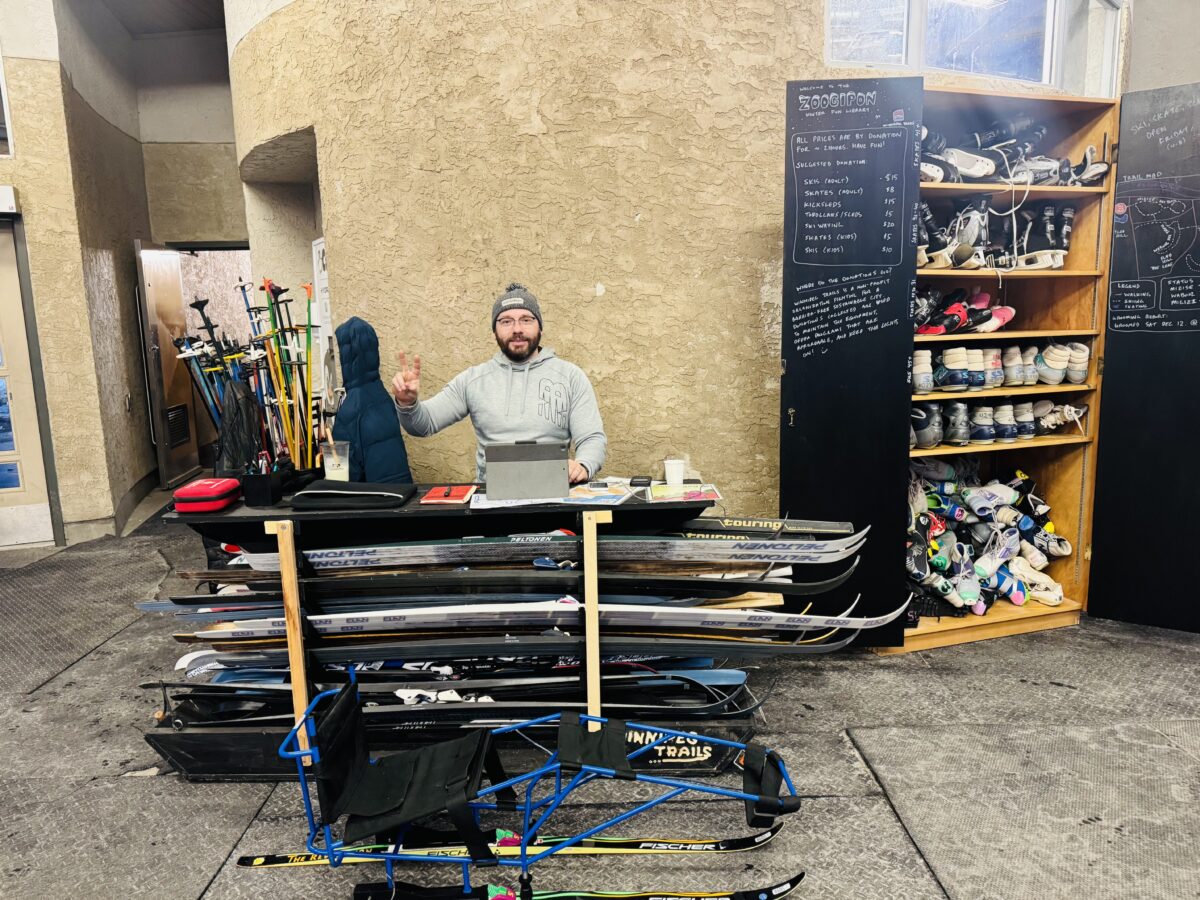 white man in winterwear stands next to skis, skates, boots, sleds, and a sit ski