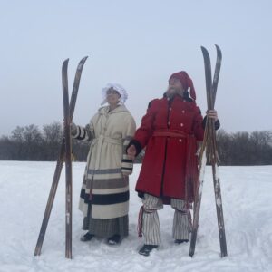 two people in period costume with long wooden skis in the snow. she is wearing a striped ribbon skirt-looking long jacket grey and white with a bonnet, he is wearing a big wollen red jacket