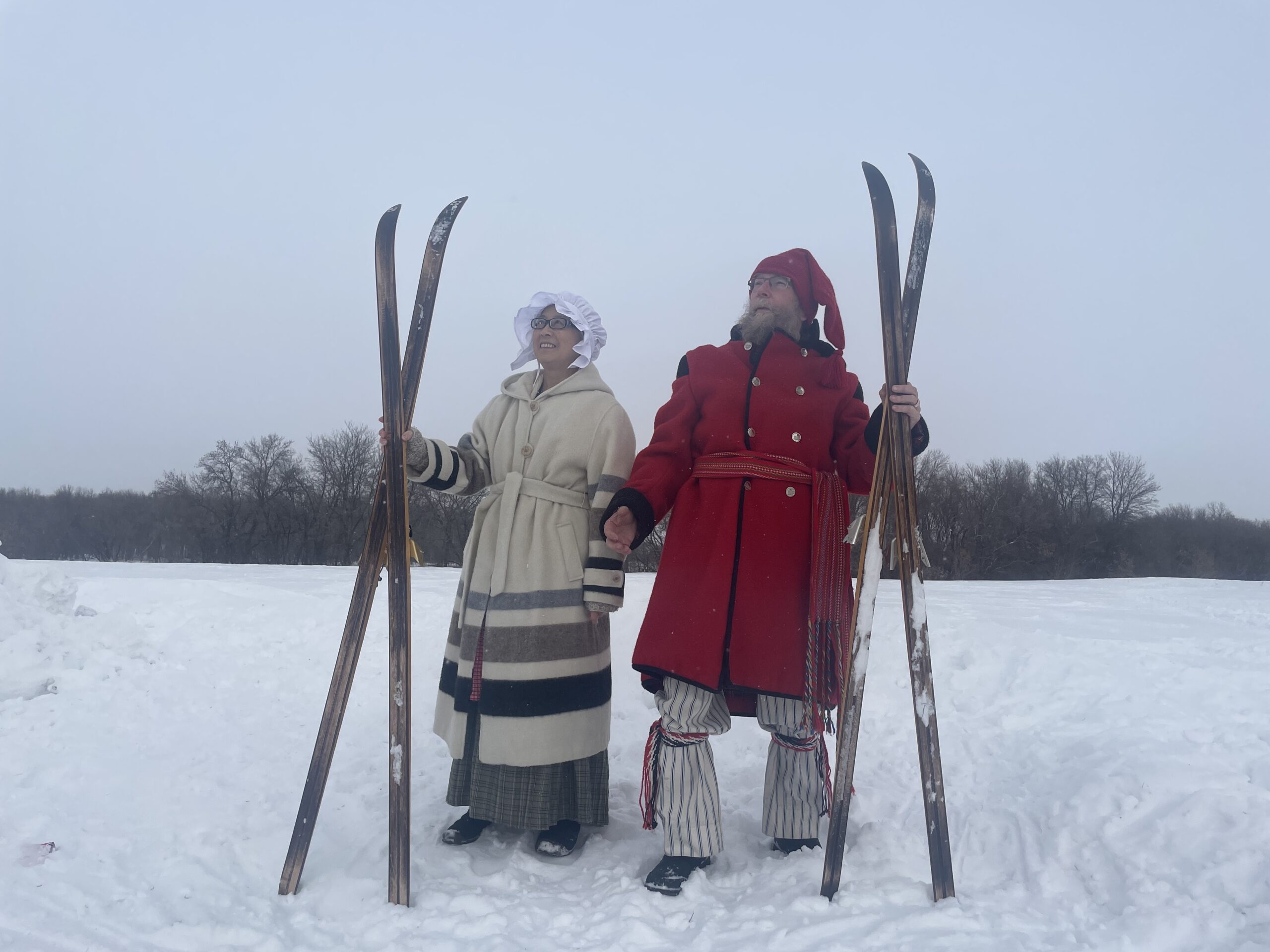 two people in period costume with long wooden skis in the snow. she is wearing a striped ribbon skirt-looking long jacket grey and white with a bonnet, he is wearing a big wollen red jacket