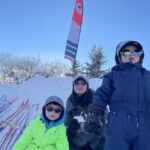 2 children and their mom wearing cool sunglasses in front of a snow pile and lots of skis