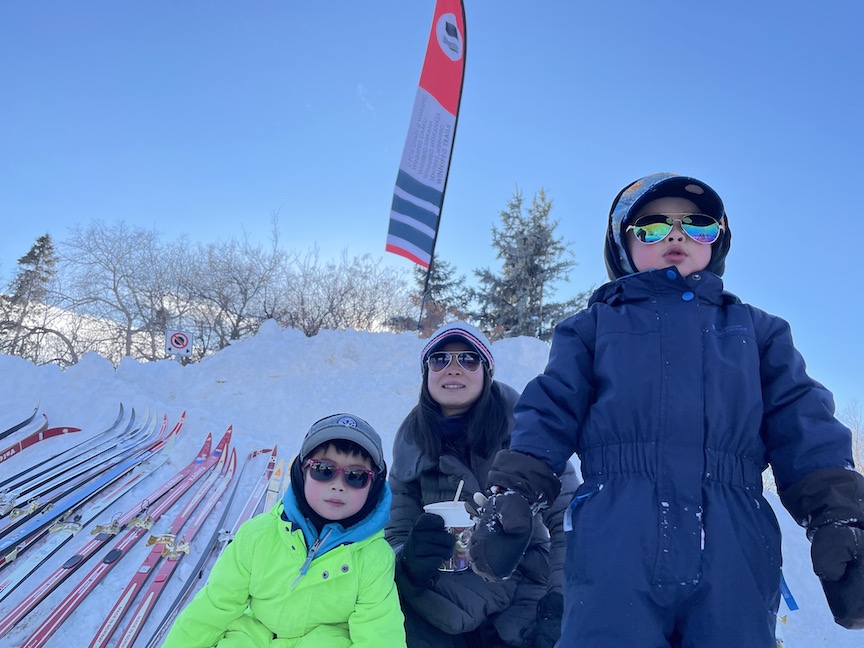 2 children and their mom wearing cool sunglasses in front of a snow pile and lots of skis