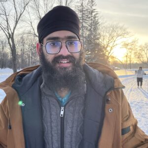 Adult man in glasses. brown jacket and black turban in front of sunset with skiier in background