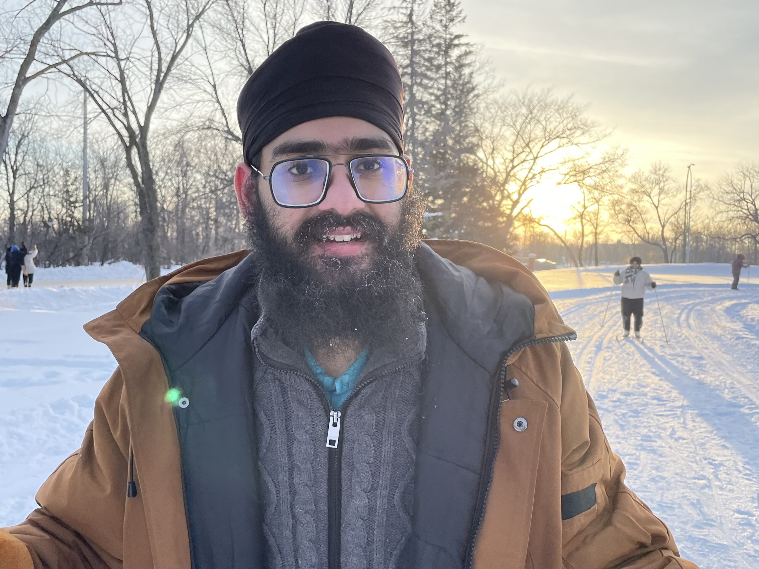 Adult man in glasses. brown jacket and black turban in front of sunset with skiier in background