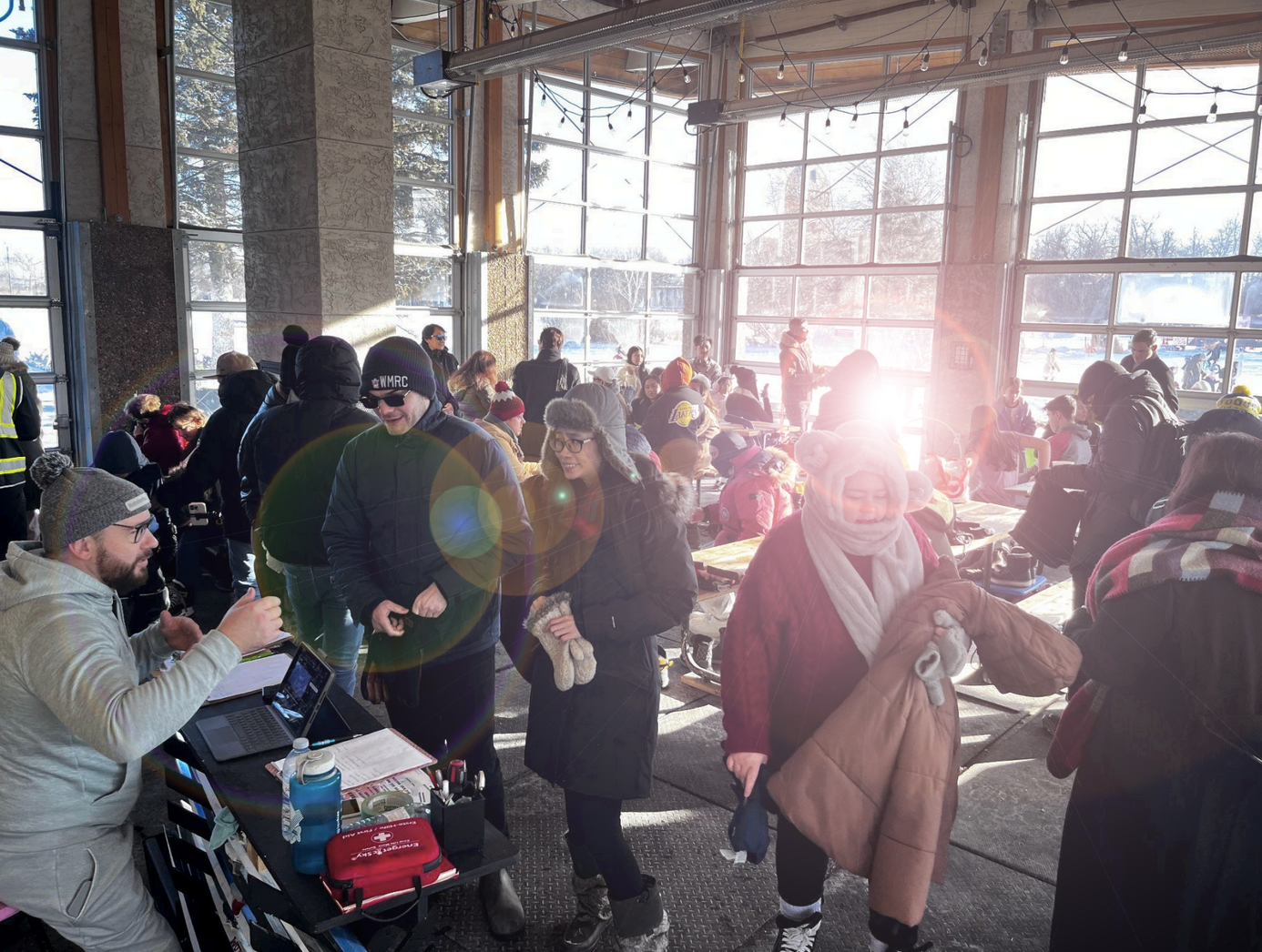 people lining up at a desk, people in the background trying on skates and skis. Sunny winter day and landscape out large overhead door windows.