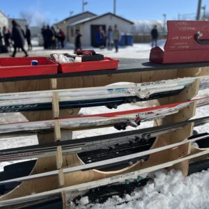 A plywood ski carrier with red tool box in front of a winter background with some buildings a hpckey rink and a crowd of people