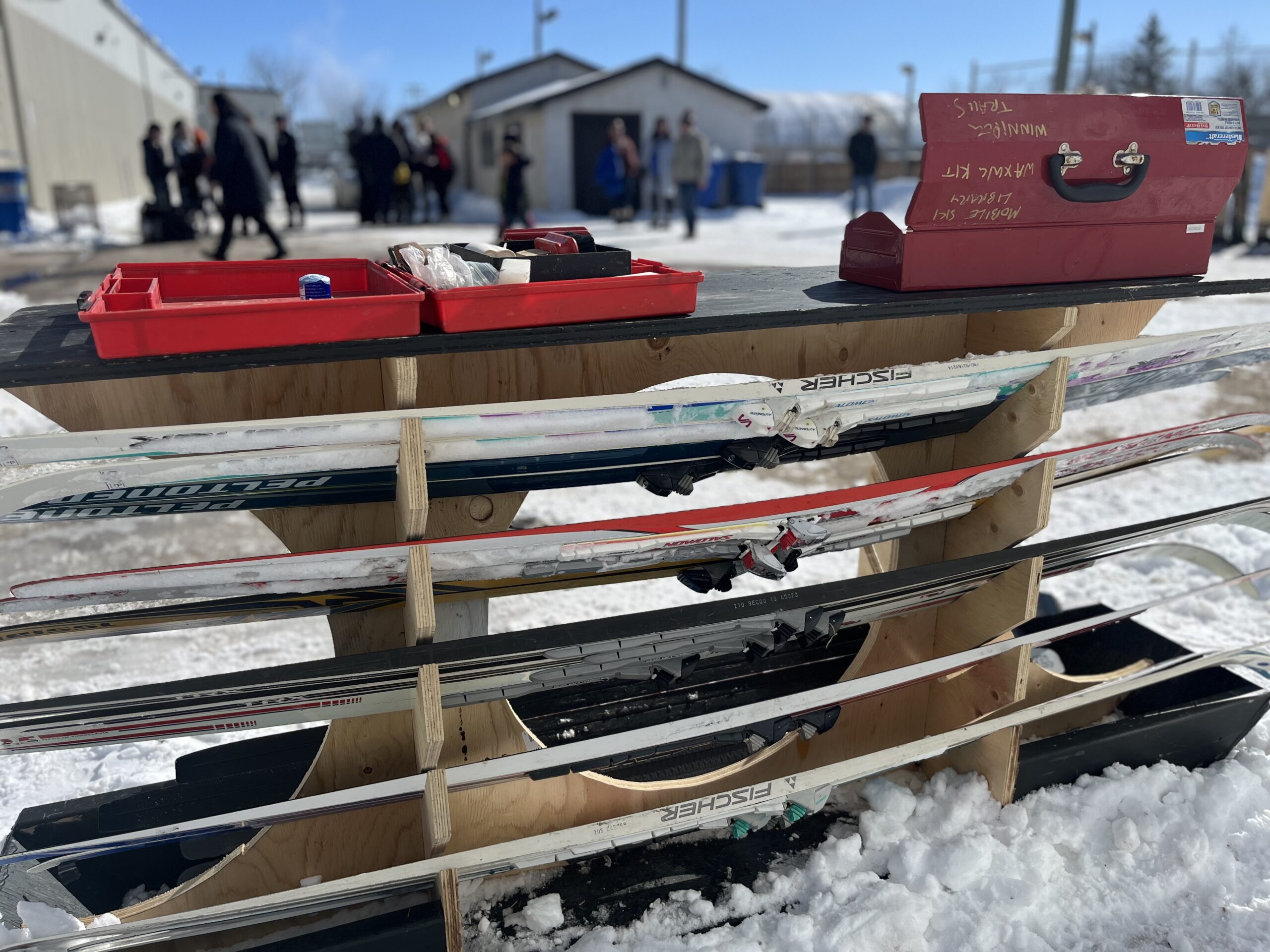 A plywood ski carrier with red tool box in front of a winter background with some buildings a hpckey rink and a crowd of people