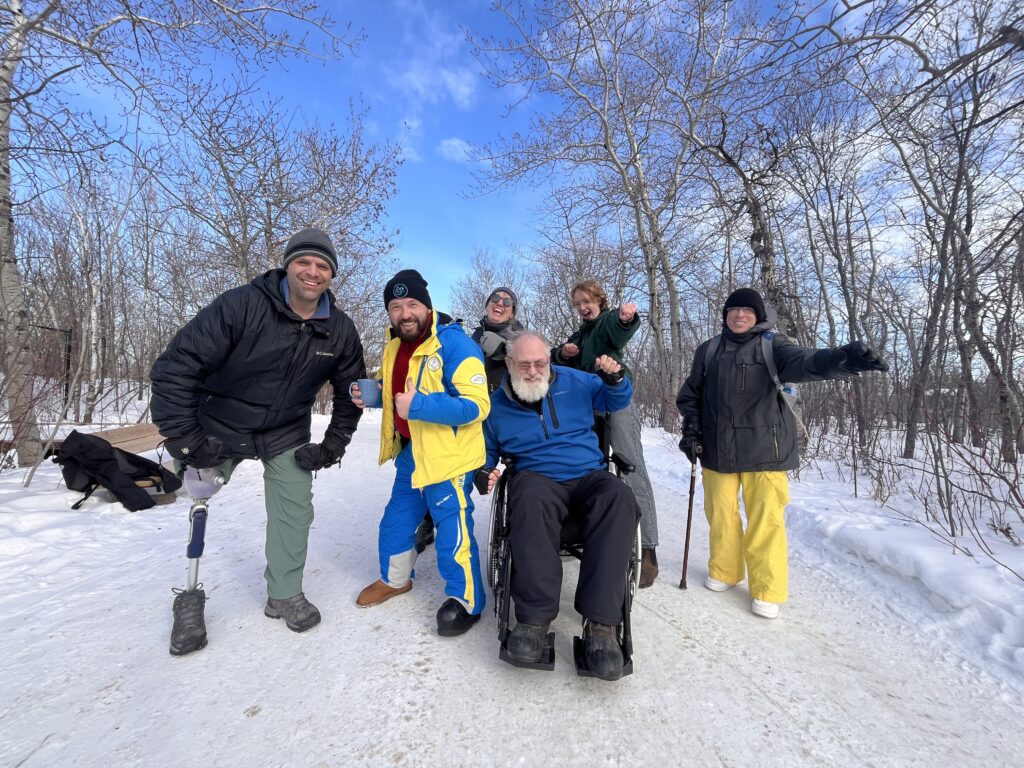 A "band photo" like picture of a group of people posing many of who have a visible diusability such as a prosthetic leg or using a mobility aid, the backdrop shows a snowy winter day in the forest and people are smiling.