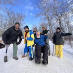 A "band photo" like picture of a group of people posing many of who have a visible diusability such as a prosthetic leg or using a mobility aid, the backdrop shows a snowy winter day in the forest and people are smiling.