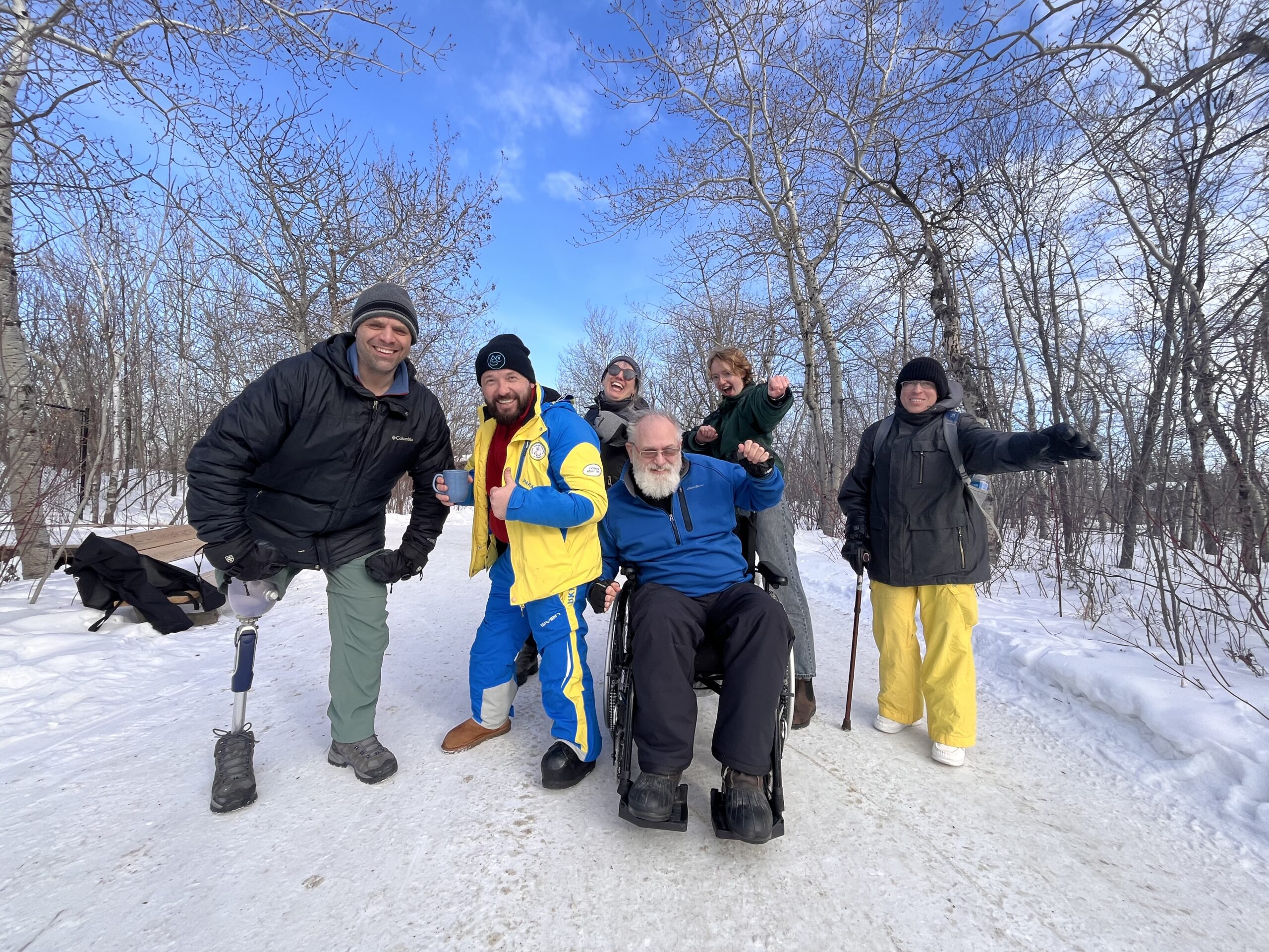 A "band photo" like picture of a group of people posing many of who have a visible diusability such as a prosthetic leg or using a mobility aid, the backdrop shows a snowy winter day in the forest and people are smiling.